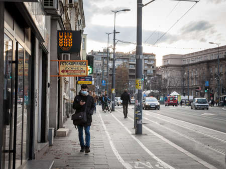 BELGRADE, SERBIA - NOVEMBER 28, 2021: Selective blur a young man, walking checking his smartphone wearing a face mask in the streets of Belgrade on coronavirus covid 19 crisis.のeditorial素材