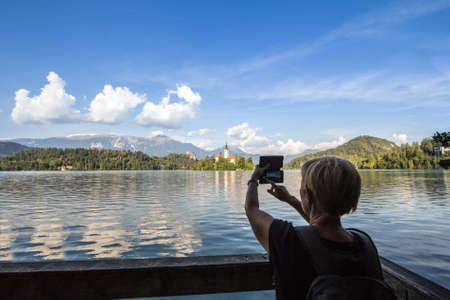 BLED, SLOVENIA - SEPTEMBER 11, 2021: Woman, a tourist, taking a photo picture of the Bled lake with her Smartphone telephone. Lake Bled, or Blejsko jezero, is a lake in Julian alps, Slovenia.のeditorial素材