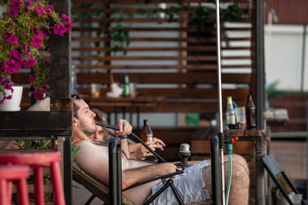 BELGRADE, SERBIA - JULY 31, 2021: Man lounging relaxing on bathing suit, smoking hookah shisha...