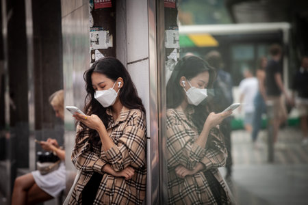 LJUBLJANA, SLOVENIA - SEPTEMBER 15, 2022: Young Asian woman, wearing a respiratory face mask checking apps on her smartphone in the street of Ljubljana, during coronavirus covid 19 health crisis.のeditorial素材