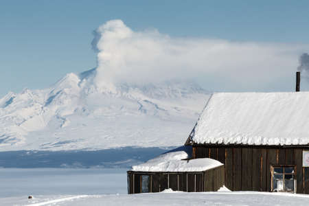 KAMCHATKA PENINSULA, RUSSIA - JANUARY 05, 2016: Winter view of the field wooden house Kamchatka volcanologists on the background eruption active Shiveluch Volcano. Eurasia, Russian Far East, Kamchatsky Krai.のeditorial素材