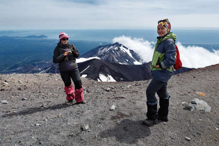 AVACHA VOLCANO, KAMCHATKA PENINSULA, RUSSIA - AUG 7, 2014: Two young women tourists stand of top of crater of active Avachinsky Volcano on background of cone of Kozelsky Volcano and Pacific Ocean.のeditorial素材