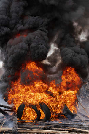 Burning cars tires, strong flame of red-orange fire and clouds of black smoke in sky. Selective focus, blur from strong fire.の写真素材