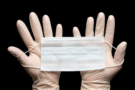 Doctor holding medical face mask in two hands in white gloves isolated on black background. Concept...