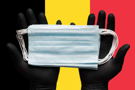 Doctor holds medical face mask, respiratory bandage in hands in black gloves on background flag of Belgium. Concept global impact of COVID-19, coronavirus quarantine, hygiene, flu, pandemic outbreak.の写真素材