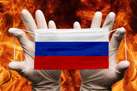 Doctor holding in white gloves protection medical face mask, respiratory bandage with Russia national country flag superimposed on mask. Concept on background of dangerous red flames natural fireの写真素材