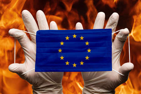 Doctor holding in white gloves protection medical face mask, respiratory bandage with European Union EU flag superimposed on mask. Concept pandemic outbreak on background of dangerous red flames fireの写真素材