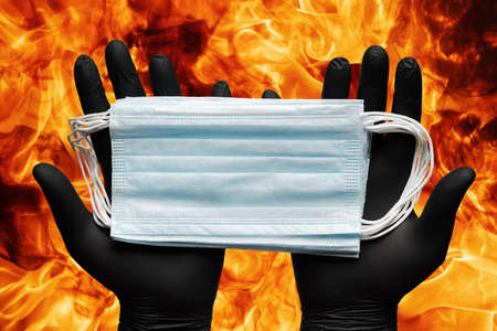 Doctor holding lot medical face masks in hands in black protective gloves on background flames of strong red fire. Concept quarantine, grippe, pandemic outbreak, hygiene, airborne diseases, flu.の写真素材