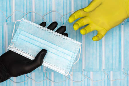 Doctor holds surgical face mask in hand in black medical glove and gives it to patient in yellow protective glove. Medicinal concept on background of lot blue respiratory bandages for human face.の写真素材