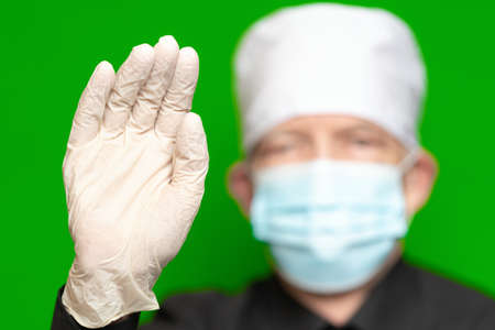 Doctor male in surgical face mask on defocus foreground put forward palm of his hand in protective latex glove foreground, waves by his hand, hello or stop sign. Concept of protection, safety on greenの写真素材