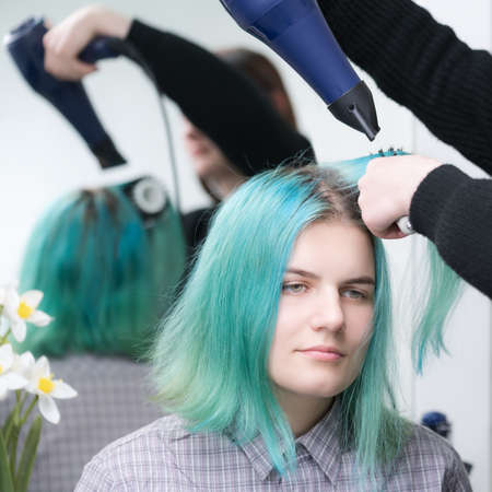 Portrait of beautiful young woman with long green hair while drying and styling hair in beauty salon. Brunette hairstylist dries customer hair with blue hairdryer and hairbrush.の写真素材