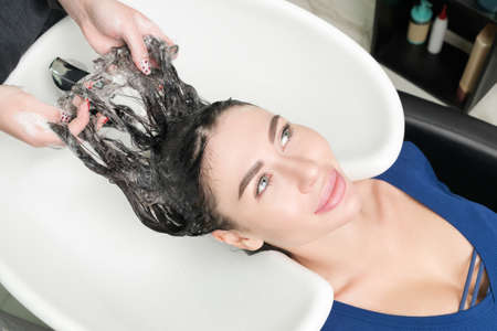 Hairstylists hands wash long hair of Caucasian brunette woman with shampoo in professional sink for...