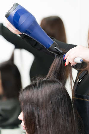 Hands of professional beautician drying long brunette hair of client using blue hairdryer and hairbrush in beauty salon.の写真素材