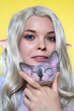 Portrait of Young blonde woman cosplay elf in blue dress, holding Sphinx cat, smiling, looking at...
