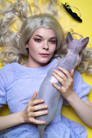 Young female cosplay elf in blue dress looking at camera with eyes of different colors, lying down on yellow background, spreading her blonde curly long hair over it. Sphinx cat lies on her chest.の写真素材