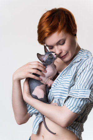 Redhead young woman with closed eyes gently hugs cat looking at owner. Studio shot on white background. Portrait beautiful hipster with short hair dressed in striped white-blue shirt. Part of series.の写真素材