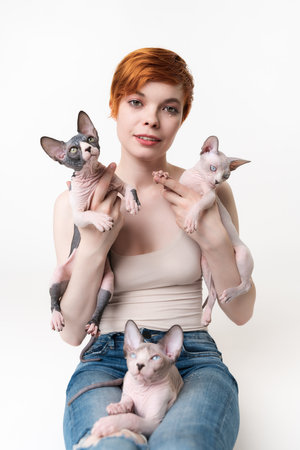 Redhead young woman holding two Sphynx Cat in hands and one kitten lying on her legs. Pretty woman with short hair in T-shirt and jeans looking camera, lying down on white background. Part of seriesの写真素材