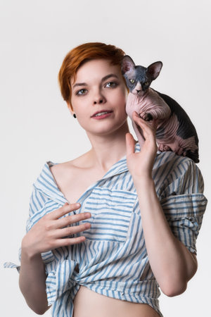Sphynx kitten sitting on shoulder of beautiful redhead young woman 25 years old. Portrait of female with short hair dressed in striped white-blue shirt. Studio shot on white background. Part of seriesの写真素材