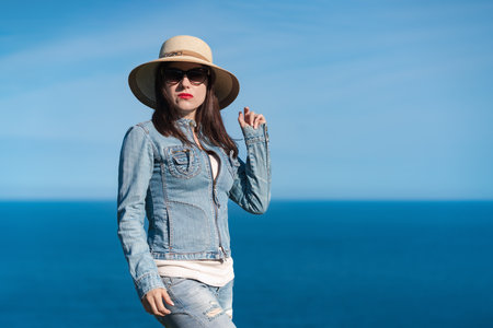 Portrait of woman in denim jacket, jeans, summer straw hat on head and sunglasses looking at camera....