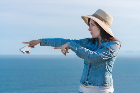 Hipster woman in straw hat and denim jacket shows with two hands direction to side. A serious 40...