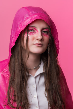 Portrait informal young adult woman with pink stage makeup with sequins and arrows painted on face, looking at camera. Female dressed in pink jacket with hooded over. Studio shot on colored backgroundの写真素材
