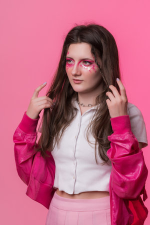 Portrait of young adult woman with pink stage make-up with sequins and arrows painted on face, dressed in pink jacket, skirt and white t-shirt. Studio shot of informal female on colored backgroundの写真素材
