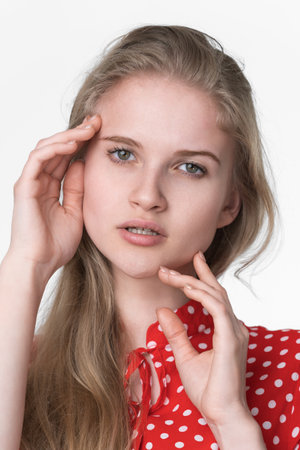 Portrait of beautiful woman face. Fashionable young blonde model with long hair dressed in summer red polka dot dress poses on white background. Elegant Caucasian female 21 years old looking at cameraの写真素材
