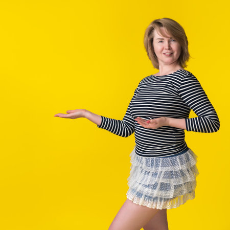 Portrait of curvy blonde woman in mini skirt and striped long sleeve shirt, pointing with open palms to space for future advertising. Woman smiling, looking at camera on yellow background. Copy spaceの写真素材