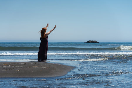 Rear view of happy redhead woman in dark purple long dress standing on summer sandy beach with raised arms reaching out to blue sky and sea waves. Full length view of barefoot female. Copy spaceの写真素材