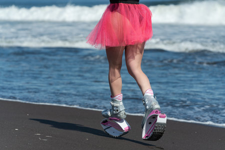 KAMCHATKA, RUSSIA - JUNE 15, 2022: Rear view of woman legs in sports boots Kangoo Jumps, black swimsuit and short skirt running on black sandy beach. Outdoors fitness workout on summer sea shoreのeditorial素材