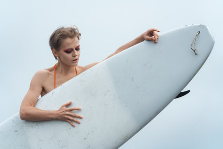 Female sports fashion model holding surfboard. She is standing behind surf board and looking down thoughtfully. Close-up view of beautiful woman surferの写真素材