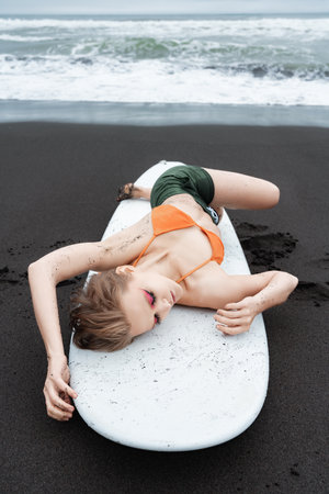 Female surfer is lying on white surfboard on black sandy beach in Pacific Ocean. With her eyes...