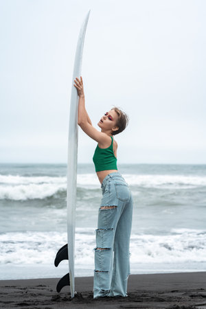 Side view of woman surfer arms raised and holding surfboard vertically. Full length playful female standing on sandy beach, posing on background of ocean waves. Hipster woman wearing casual clothingの写真素材