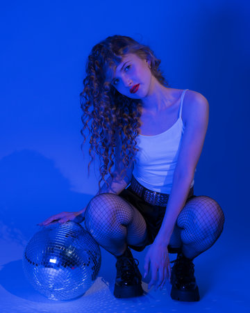 Woman with disco ball posing in squatting position while partying in nightclub. She exudes...