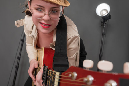 Cheerful female guitarist with glasses and grin on face is holding guitar and pointing it at camera. Playful expression musician is having fun, enjoying playing guitar during performance on stageの写真素材