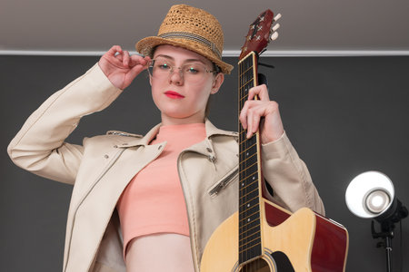 Portrait of young female guitarist holding acoustic guitar in one hand, adjusts her eyeglasses with other hand and looking confidently at camera. Musician is dressed in beige leather jacket, straw hatの写真素材