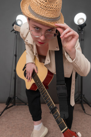 Female guitarist in glasses holds guitar and confidently looks at camera, bowing to camera and beautifully bending while holding guitar. With one hand she holds brim of straw hat on her headの写真素材