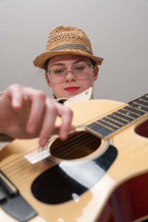 Low angle view of face young female guitarist with glasses and straw hat playing acoustic guitar and looking at camera. Dynamic perspective creating an exuberant, lively atmosphere during performanceの写真素材
