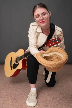 Cheerful woman guitarist with acoustic guitar in one hand and straw hat in other, stretching forward. Woman standing on one kneeの写真素材