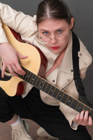 Young female hipster guitarist with glasses and straw hat playing six string acoustic guitar and looking at camera. Cropped view of playful woman musician in beige leather jacket stands on one kneeの写真素材