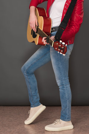 Side view of lower half of an unrecognizable female guitarist playing six string acoustic guitar. Stylish youthful musician is wearing in red leather jacket, blue jeans and sneakersの写真素材