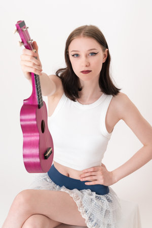 Female ukulele artist is holding instrument in her hand and extending her arm forward. Her body is oriented in seated position and she is facing camera. Musician is wearing white tank top, short skirtの写真素材