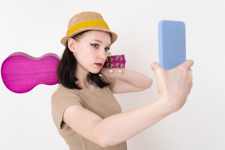 Young female musician takes selfie with mobile phone while ukulele slung over her shoulder. Stylish 18 year old Caucasian woman ukulelist poses with her cell phone held outstretched in front of herの写真素材