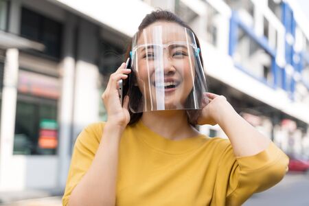 Asian woman is holding a phone outside, wearing a mask or face shield to protect against germs and viruses. Dust prevention and prevention of toxic fumesの写真素材