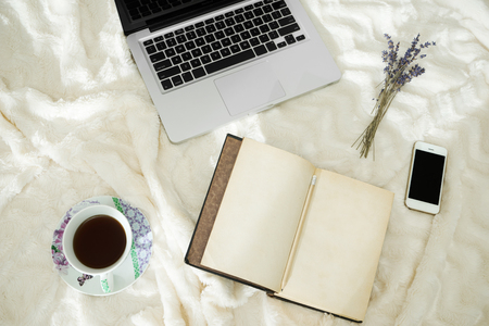 Cup of coffee, laptop, smartphone, vintage notebook and dry lavender in bed, morning lightの写真素材