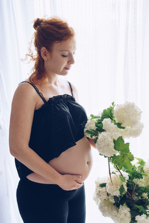 Young beautiful pregnant woman standing near window at homeの写真素材