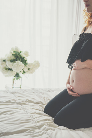 Pregnant woman sitting in bed and touching her belly with hands on window backgroundの写真素材
