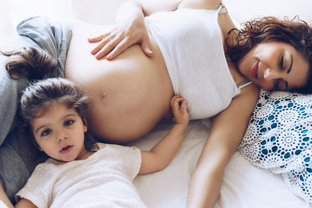 Beautiful pregnant woman and her cute little daughter together in bedの写真素材