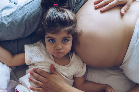 Beautiful pregnant woman and her cute little daughter together in bedの写真素材