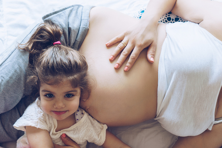 Beautiful pregnant woman and her cute little daughter together in bedの写真素材